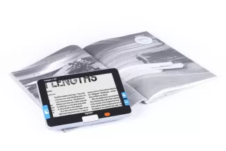 siyah-beyaz bir dergi sayfasının üzerine yerleştirilmiş Optelec Compact-7 HD taşınabilir büyüteç cihazı görülüyor. ekranında "LENGTHS" kelimesinin büyütülmüş hali yer alıyor. Cihazın kenarlarında mavi renkte büyütme seviyesinin ayarlanmasını sağlayan artı ve eksi düğmeleri var, Arka planda açık bir dergi sayfasında modern bir yapı fotoğrafı bulunmakta.